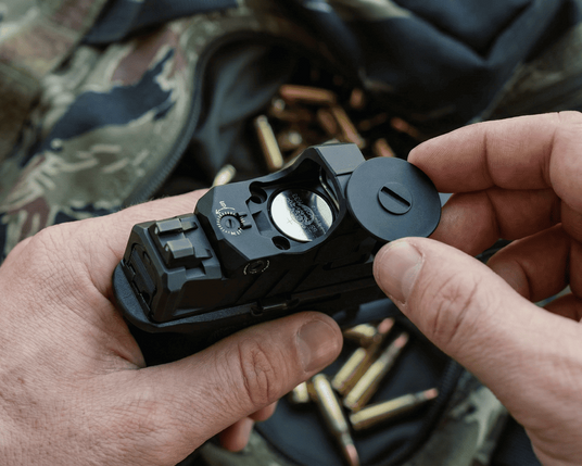 Person handling a black mercury reflex sight, changing the battery with camouflage clothing and ammunition in the background 